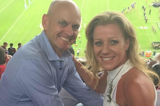 a bald man his blonde wife posing for a photo in the stands of an NRL game