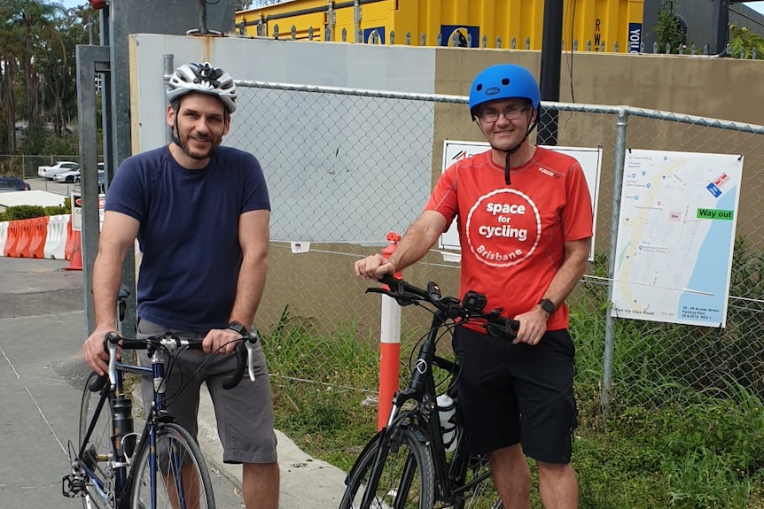 Two white men standing near bicycles, wearing helmets