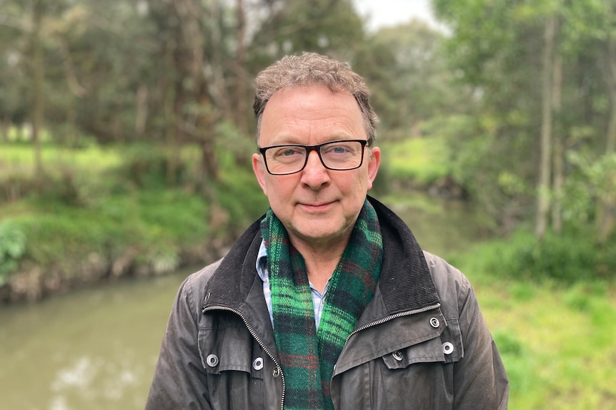 a man wearing glasses looks at the camera. There is a creek behind him. He has short hair and wears a scarf.