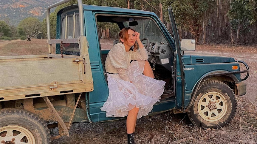Alice sits in the drivers seat of a stationary green ute with the door open. She is wearing a white dress and combat boots.