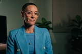Female doctor wearing blue top and jacket in an office. Her hair is close-cropped and she wears glasses.