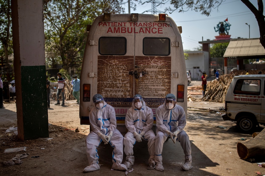 Three workers in hazmat gear sit on the back of an ambulance