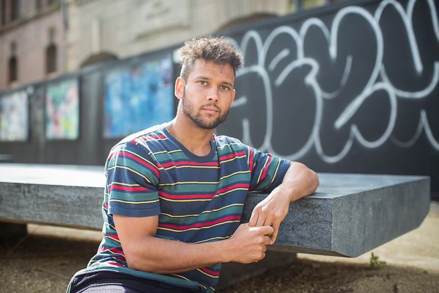 Colour photo of actor Kyle Shilling sitting against park bench on a sunny day.