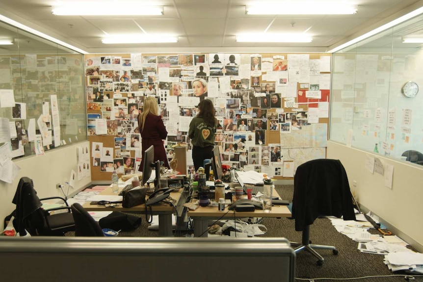 Wide shot office with two cluttered desks, paper files spread over the floor and pictures relating to case on noticeboard.