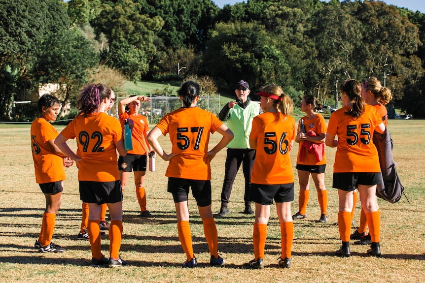 A team of soccer players wearing orange and black uniforms stand in a circle as a man in a yellow vest talks