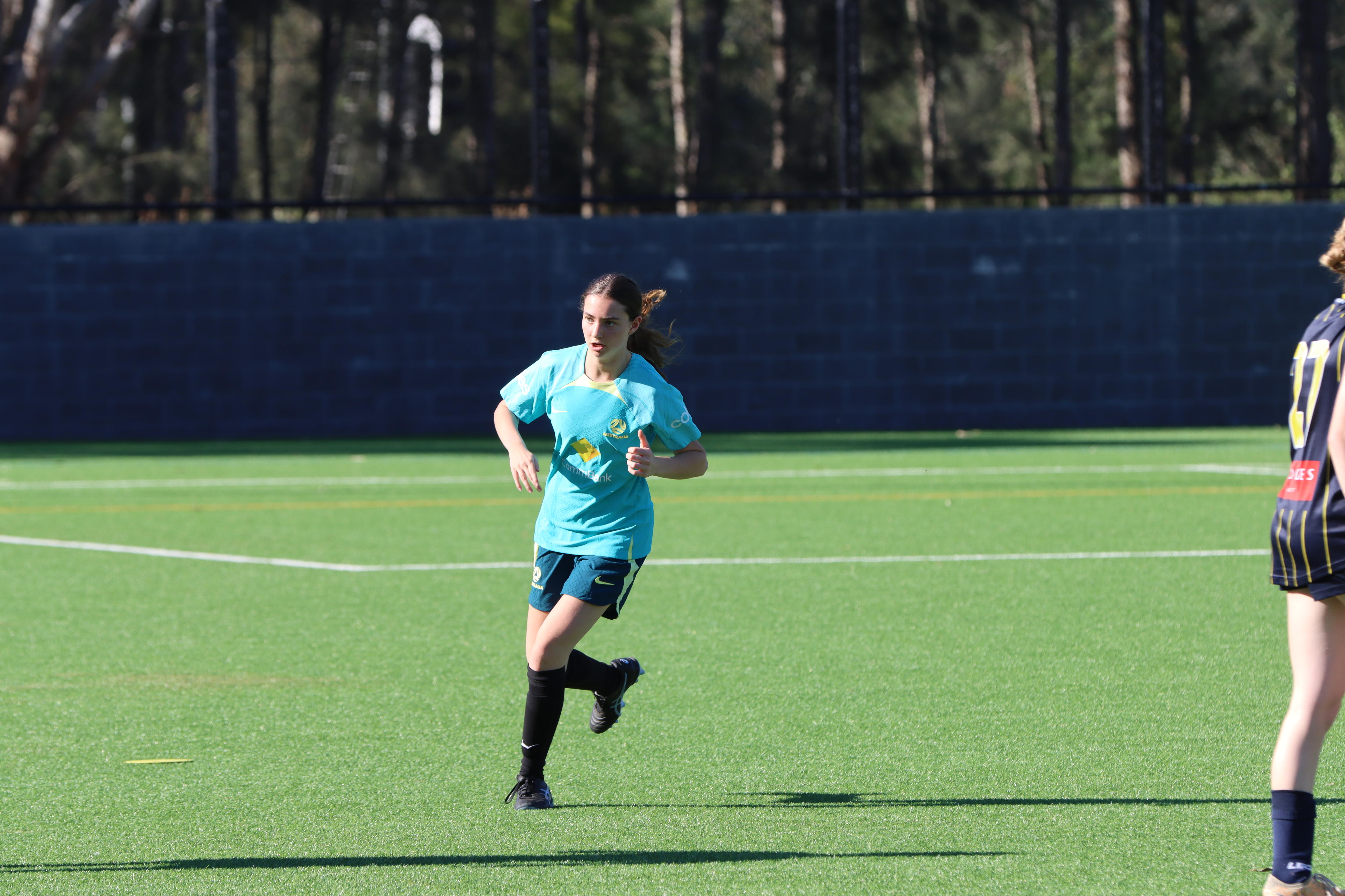 A woman wearing a turquoise soccer jersey, green shorts, black socks and shoes runs on a backdrop of green grass.