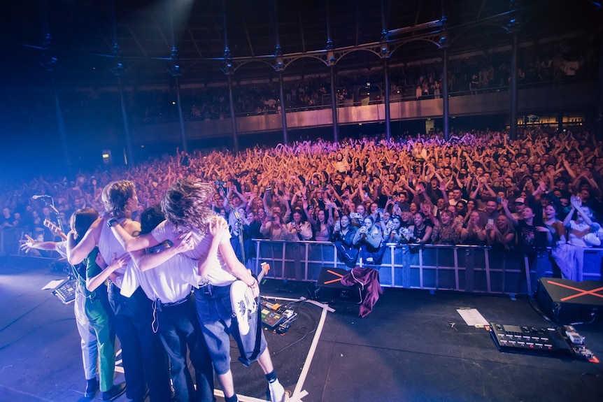 A band stands on stage with their arms around each other while they look out at a large crowd