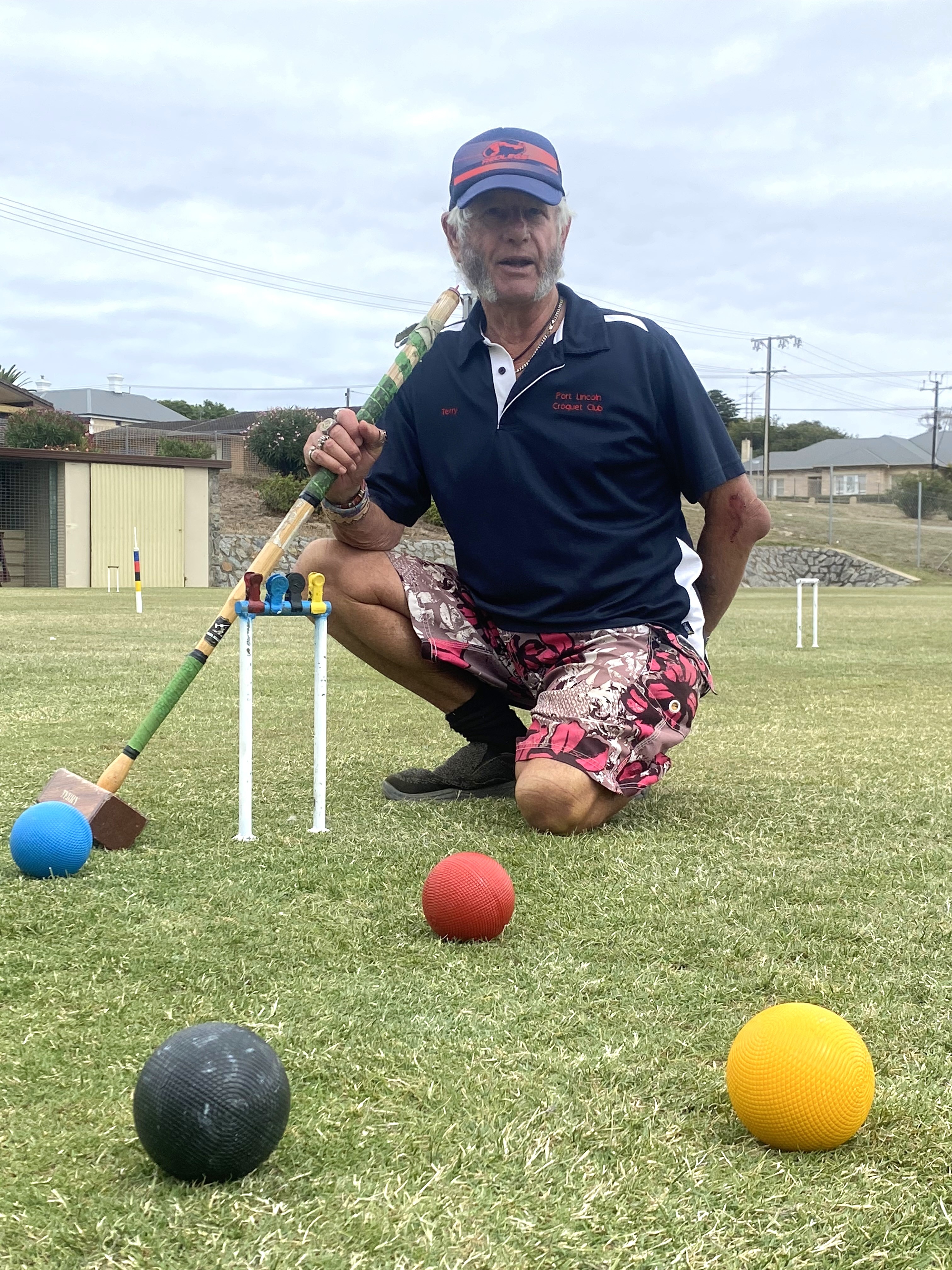 Homem de boné ajoelhado perto do aro, com macete e bola amarela, preta, azul e vermelha espalhadas na frente