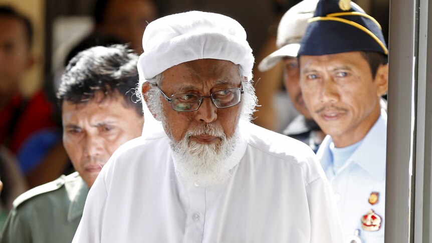 Abu Bakar Bashir walks into court, wearing all white, walks into a courtroom with police following him.