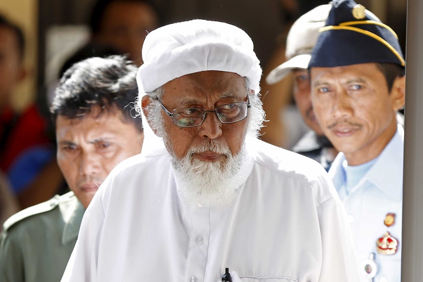 Abu Bakar Bashir walks into court, wearing all white, walks into a courtroom with police following him.