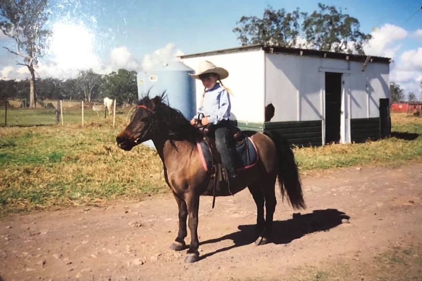 Young girl with pigtails and a cowgirl hat sits on small brown horse. 