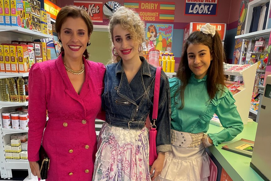 Three women in retro clothes stand inside a corner store. 