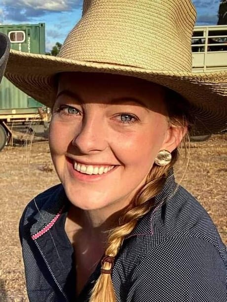 A woman with a plait on her left shoulder wearing an akubra and smiling at the camera with farm equipment behind her