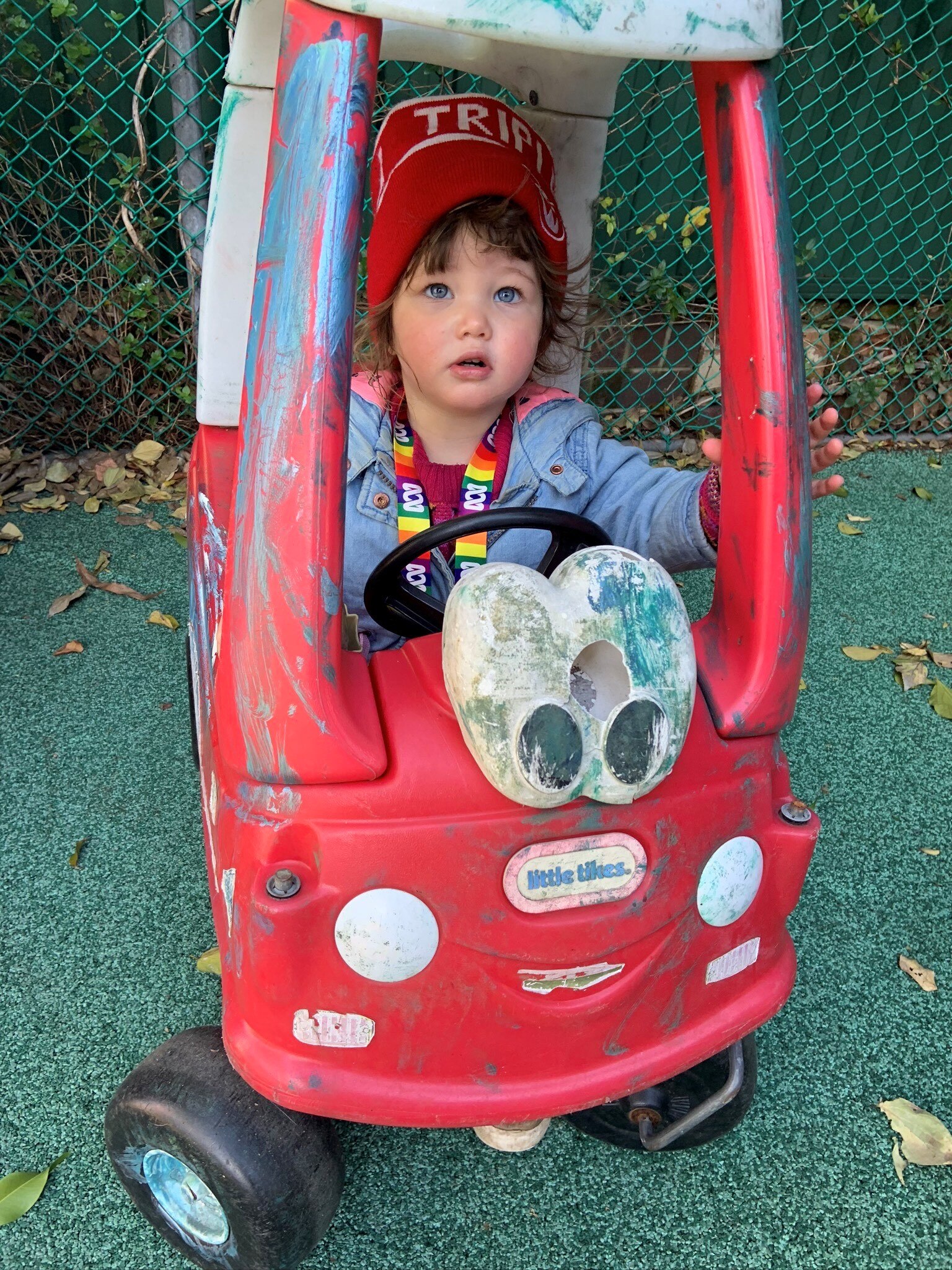 A toddler sits in a busted-up red toy car in a playground. She is wearing a triple j beanie and ABC lanyard.