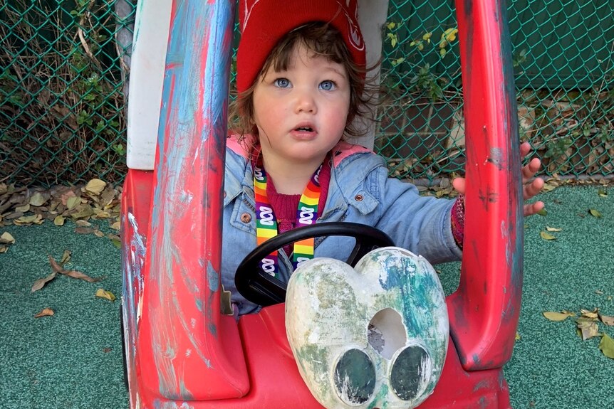 A toddler sits in a busted-up red toy car in a playground. She is wearing a triple j beanie and ABC lanyard.