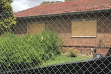 A boarded up brick building as seen through a wire fence.