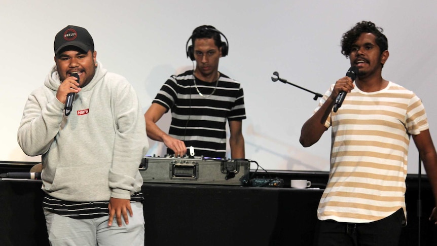 three men perform on stage with microphones and a DJ desk.