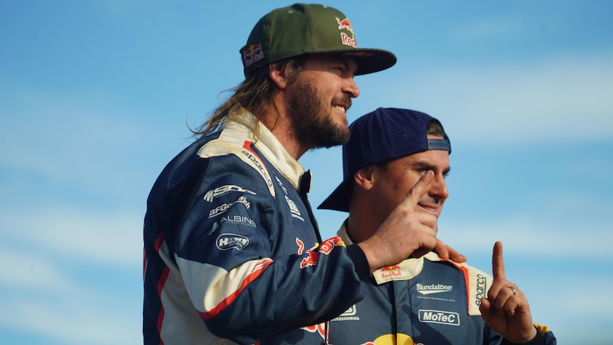 two men in car racing suits and caps gesturing 'number 1'
