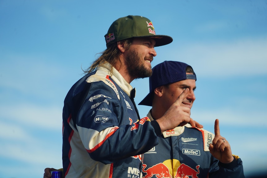 two men in car racing suits and caps gesturing 'number 1'