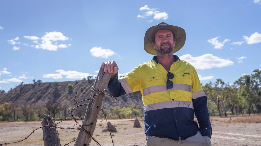 Man leans on wooden fence with barbed wire with high vis shirt, hat and sunglasses with the old mine in the background