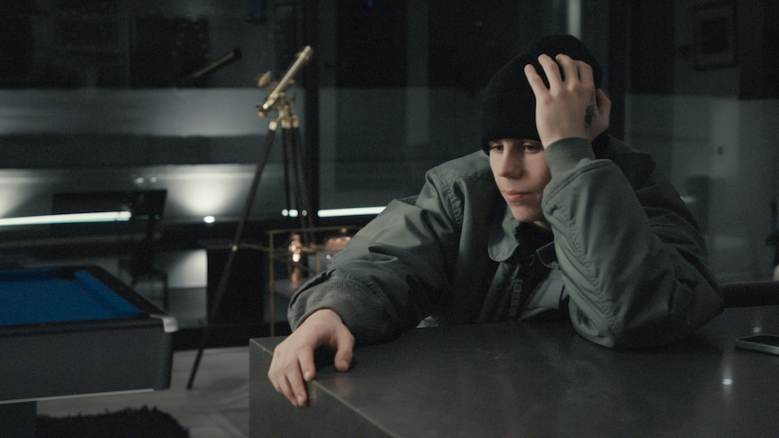 The Kid LAROI sits at a desk in a grey jumper and black beanie with his head in one hand