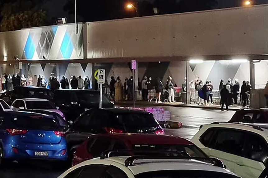 A queue of people waiting on a dark,wet  street next to parked cars. 