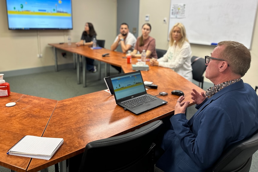 A man sits in front of a computer presenting an online program to a group of people in a meeting room 