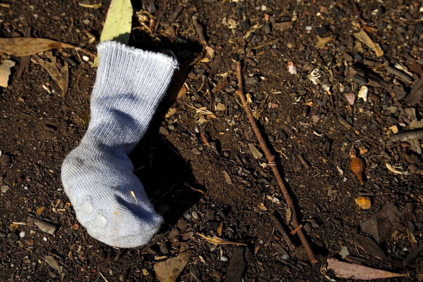 A small sock with grips at the bottom lying on the dirt, surrounded by twigs and dried leaves.