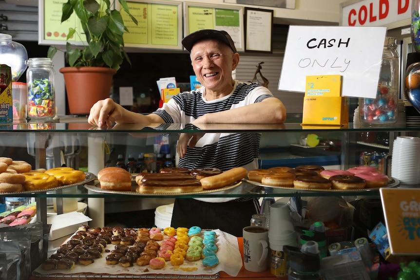 A man smiles at the front counter of his doughnut shop, filled with cinnamon, jam and iced doughnuts and eclairs.