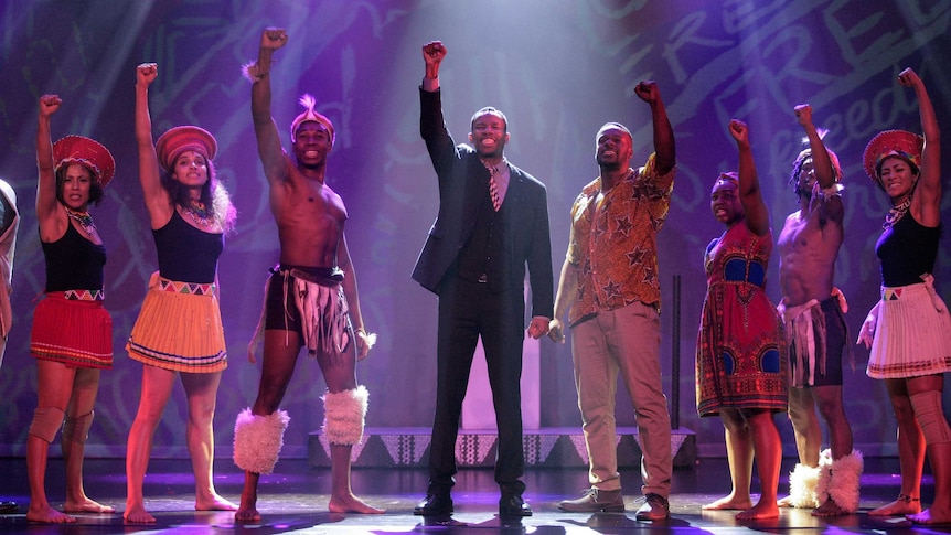 Perci Moeketsi on stage in a dark suit with a fist raised flanked by seven people in various traditional South African outfits.