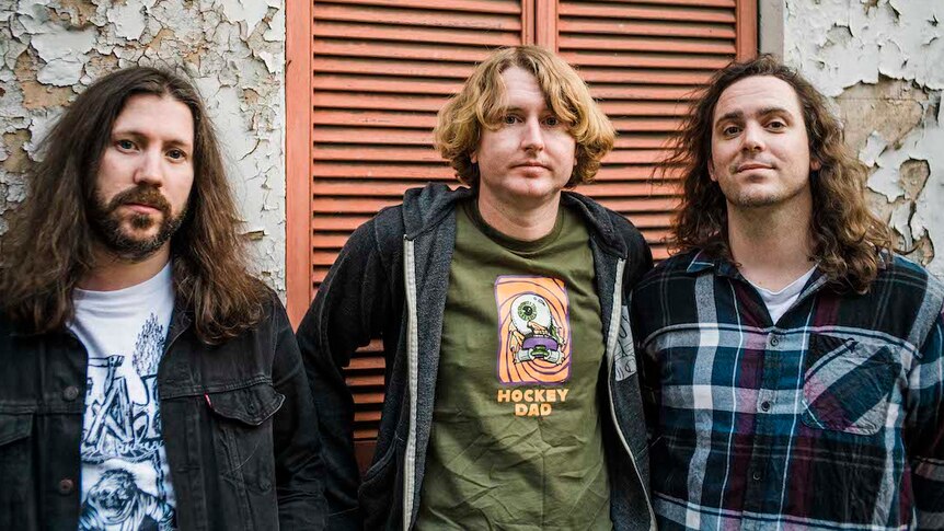 Three members of DZ Deathrays wearing band shirts, flannel shirts and jackets, standing against a brick wall and shutter blinds.