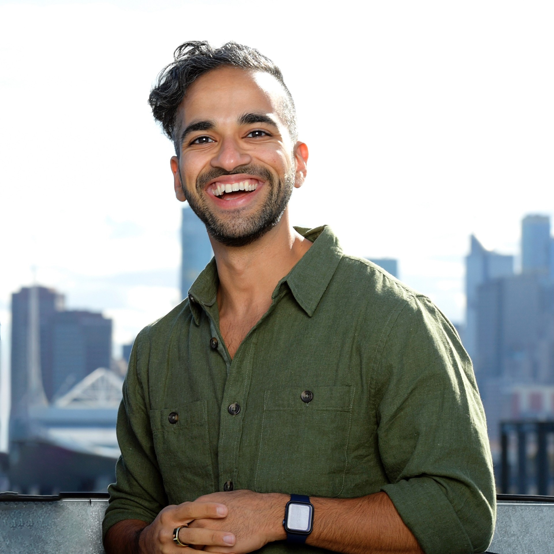 A young smiling man with dark hair and a dark green shirt standing outside.
