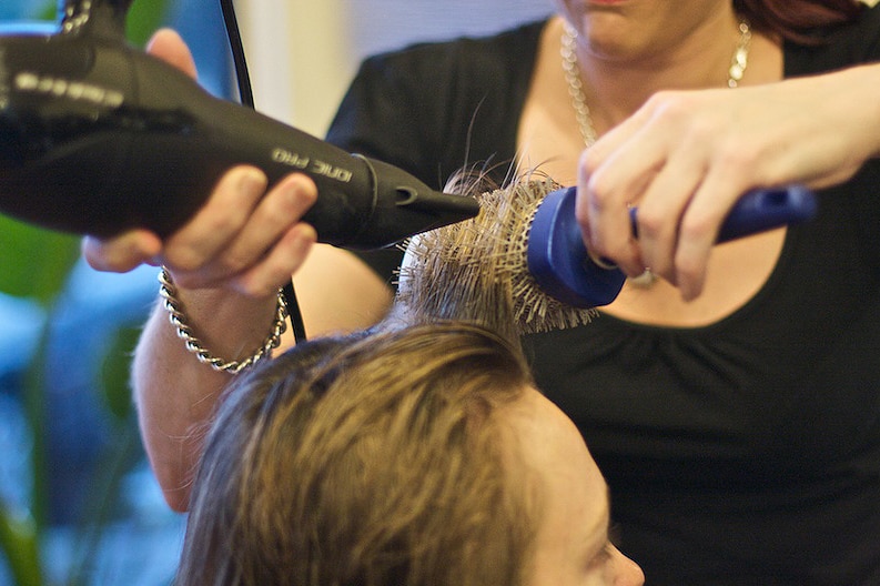 A hairdresser blow dries a customer's hair. Both faces are obscured.
