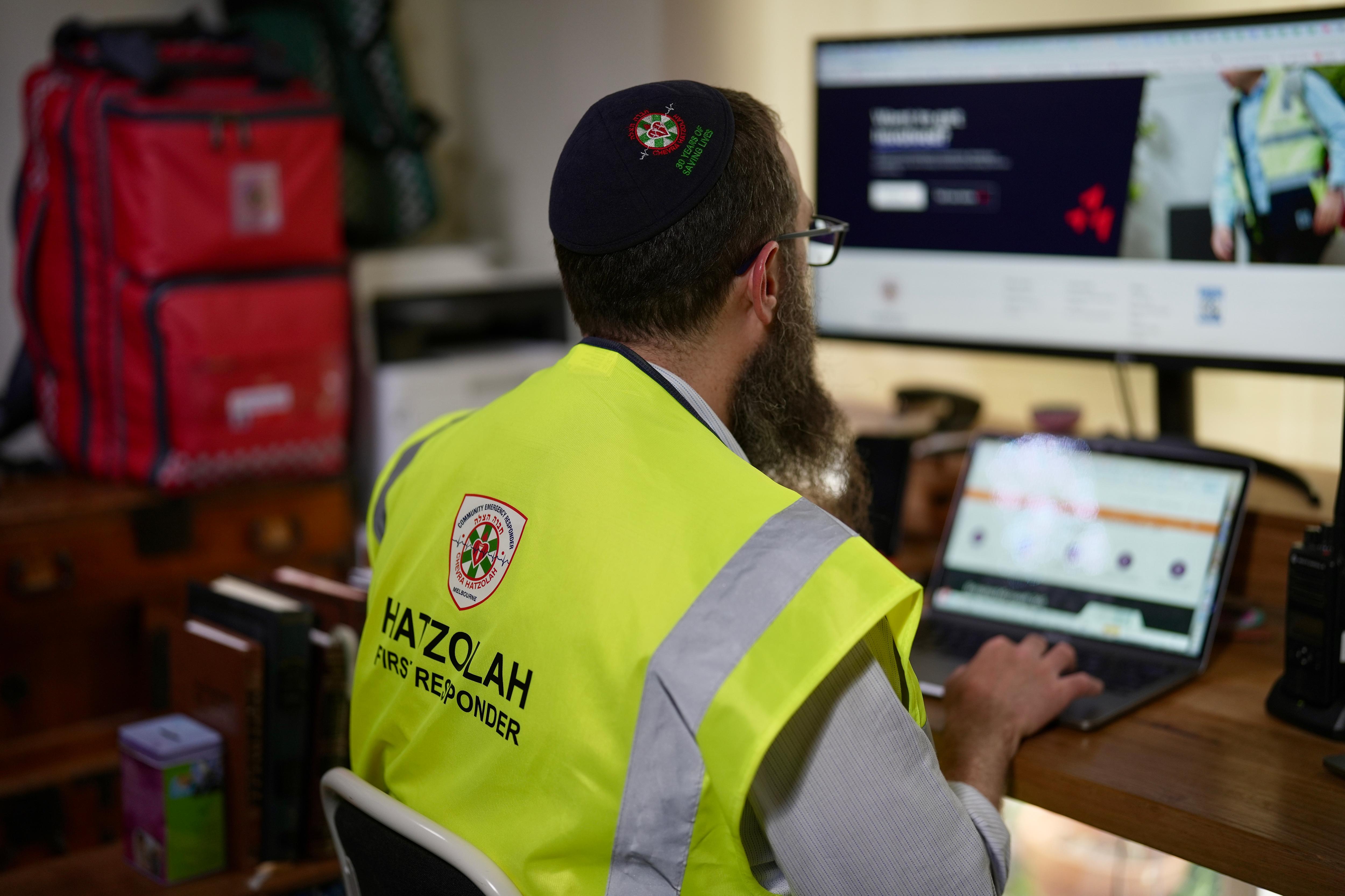 Um homem barbudo, usando óculos, solidéu e colete amarelo, ambos com o emblema do Hatzolah, está sentado em frente a um computador ao lado de uma bolsa vermelha.