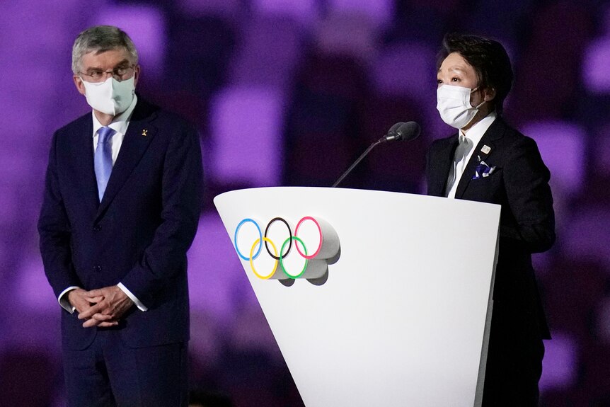 Thomas Bach bumps elbows with Seiko Hashimoto near a lectern featuring the Olympic rings. 