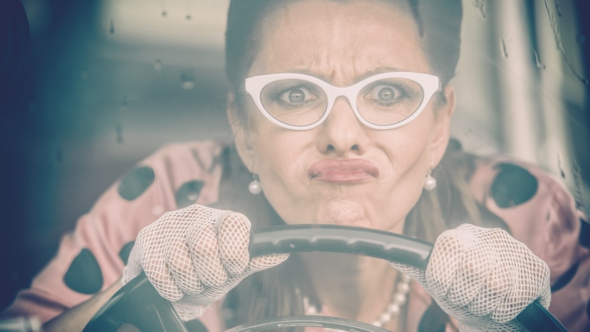 a woman wearing glasses and lace gloves behind a driving wheel pulling a disenchanted face