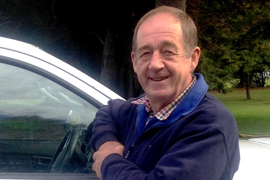 A man in a navy jumper leaning on a white vehicle.