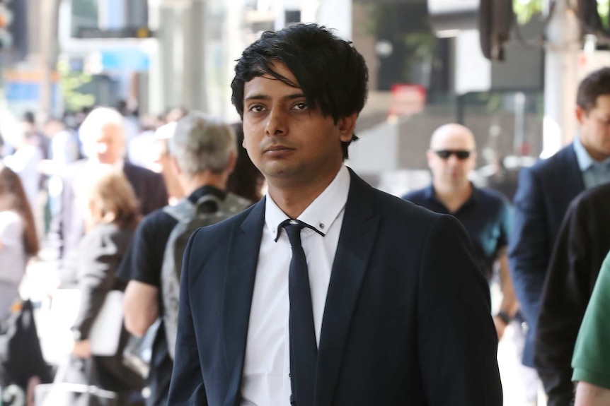 Samandeep Singh wears a black suit and tie and white shirt and is walking outside court on a sunny day.
