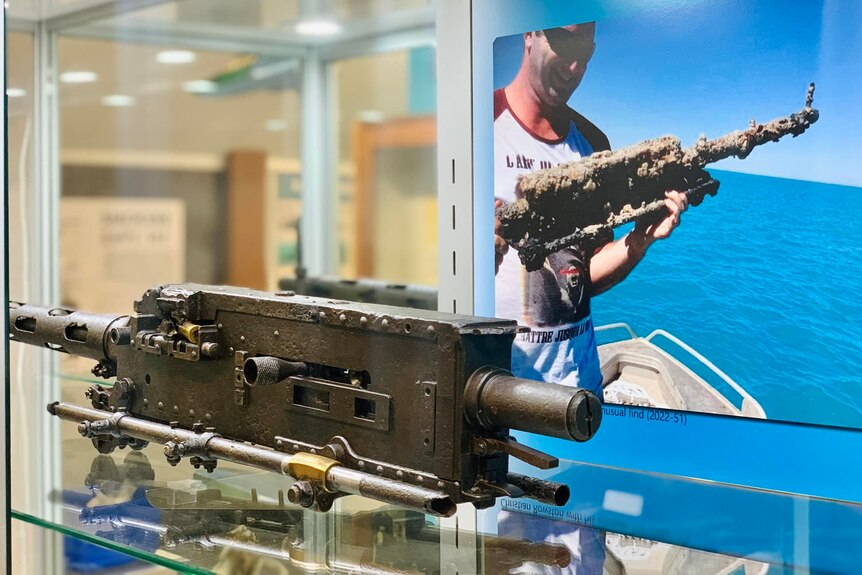 A gun in a glass case, with a photo in the background of it being pulled from the water.