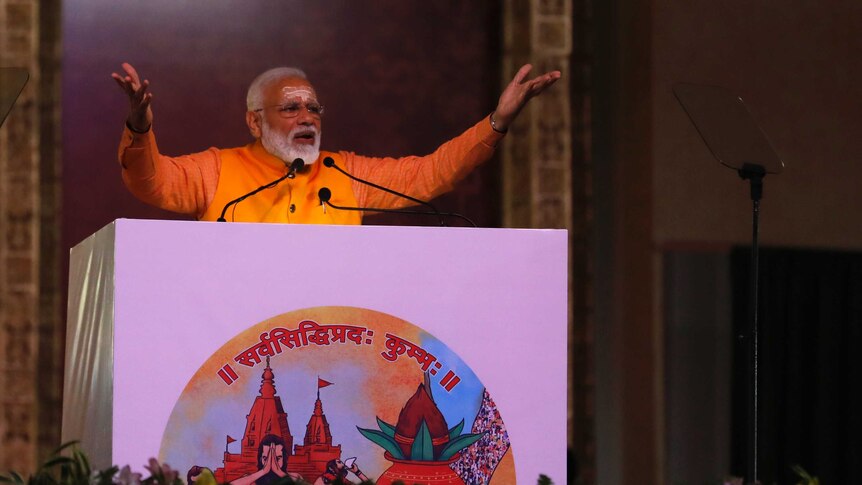 India's PM Narendra Modi holds his hands in the air,  speaks at a podium wearing an orange shirt, white script on his forehead