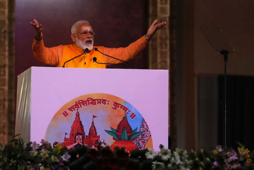 India's PM Narendra Modi holds his hands in the air,  speaks at a podium wearing an orange shirt, white script on his forehead