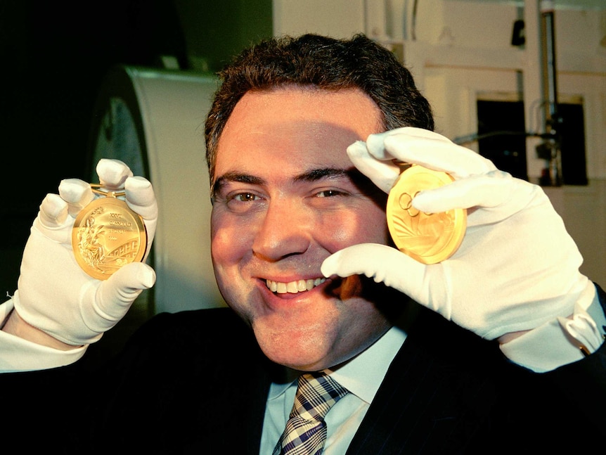 A man with brown hair wearing surgical gloves holds up two gold medals, one in each hand