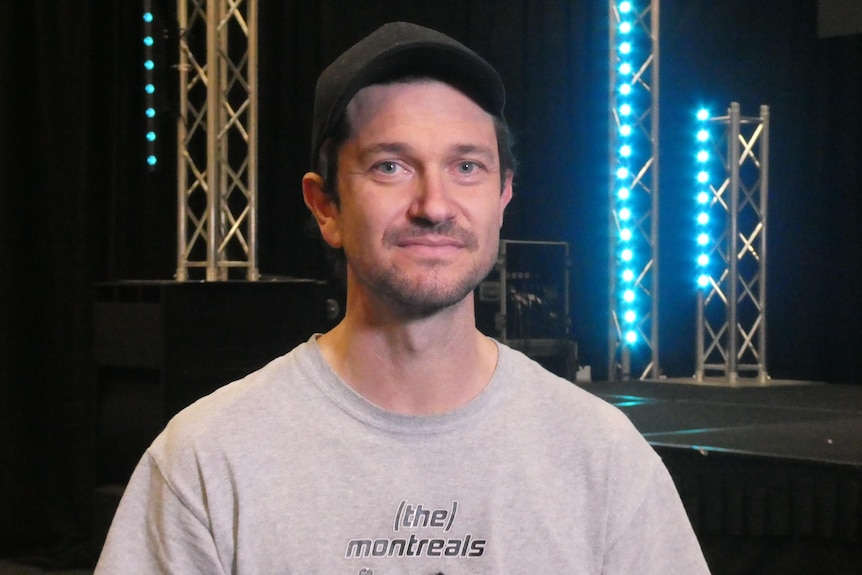 A man wearing a black cap and grey shirt smiles at the camera in front of blue LED lights