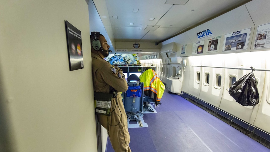 An interior scene on an aeroplane, with a man leaning against an internal wall wearing a headset and NASA jumpsuit.