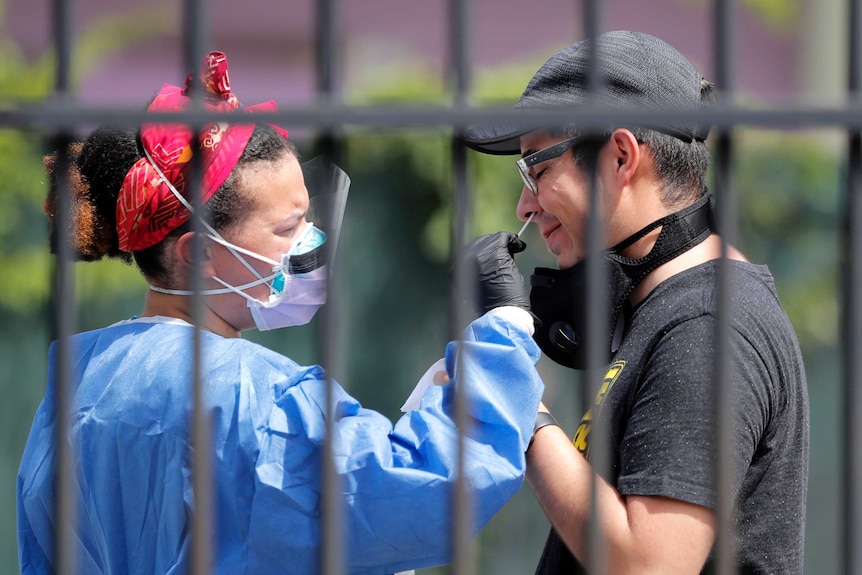 A nurse wearing protective equipment sticks a swab up the nose of a man wearing a baseball cap and black shirt.
