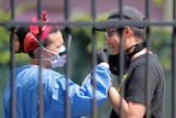 A nurse wearing protective equipment sticks a swab up the nose of a man wearing a baseball cap and black shirt.