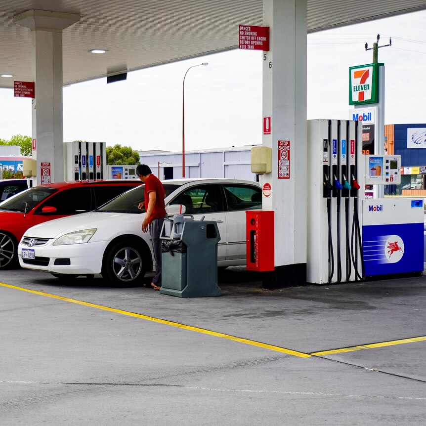 Multiple cars line up at a petrol station to pump fuel. A man in a red shirt is attending to a white car. It is overcast.
