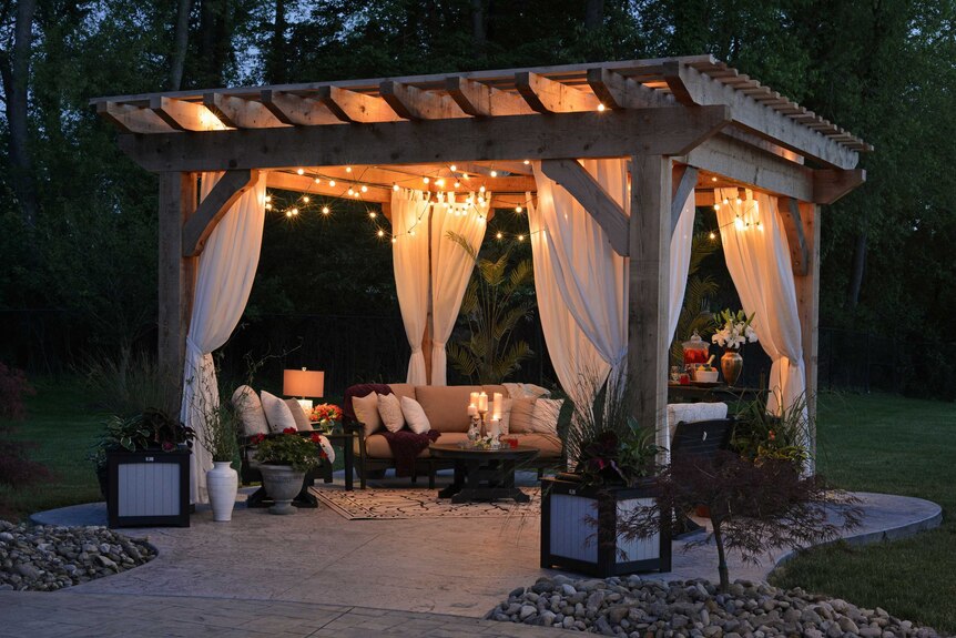 A wooden gazebo on a patio with couches, cushions, candles, plants and flowers underneath at dusk.
