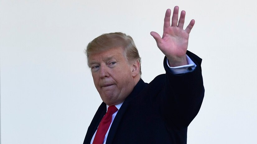 Donald Trump walks next to an outer wall of a building, with his hand raised in a wave.