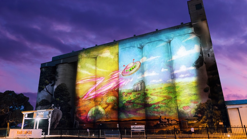 A colourful artwork of a spaceship projected on a silo under a purple darkening sky.